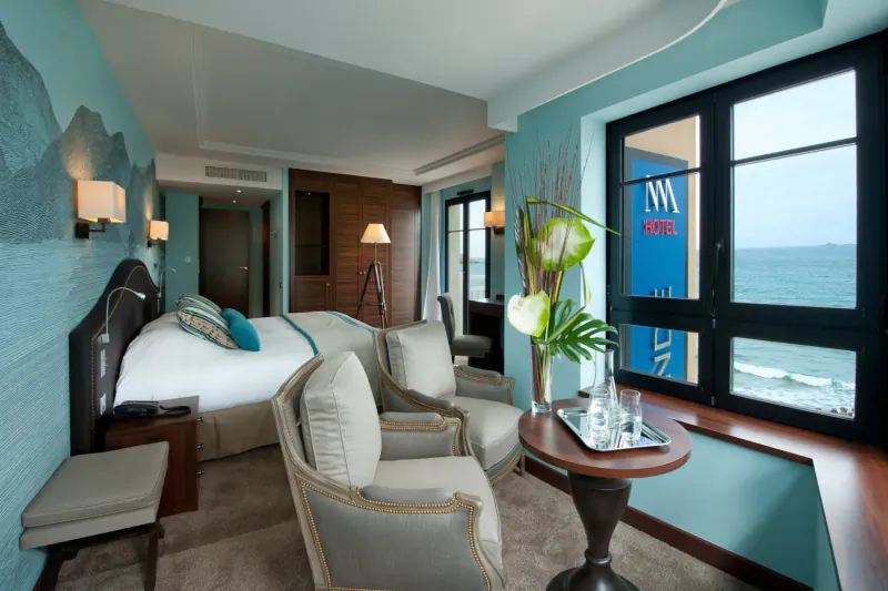 Luxurious hotel room at Nouveau Monde in Saint-Malo with king bed, armchairs, tray table, ocean view window.