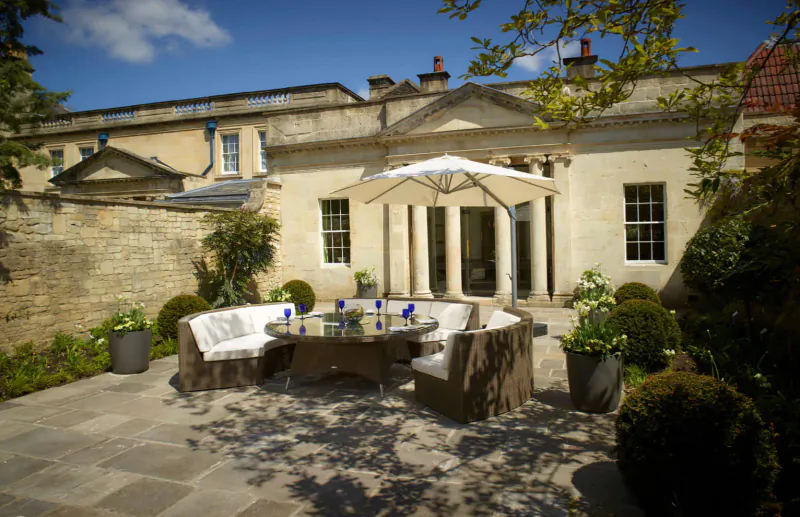 Elegant outdoor seating area with round glass table, blue glasses, and white umbrella at Royal Crescent Hotel garden party.