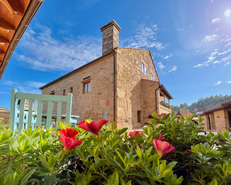 Stone house at Terranam Wellness Retreat with chimney, turquoise chair, red flowers in foreground, blue sky and hills.