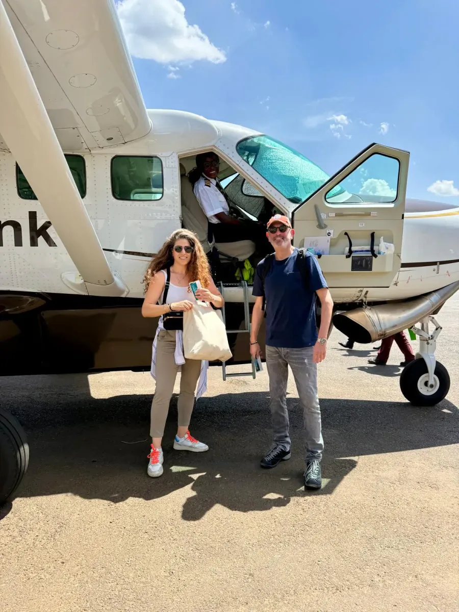 Woman in sunglasses holding tote bag and man in shades pose smiling by white Cessna airplane on sunny tarmac.