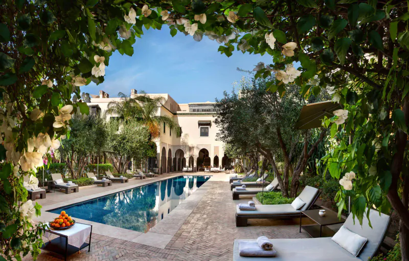 Framed by white bougainvillea arches, a luxurious Marrakech riad's poolside with lounge chairs, olive trees, and fruit bowl.