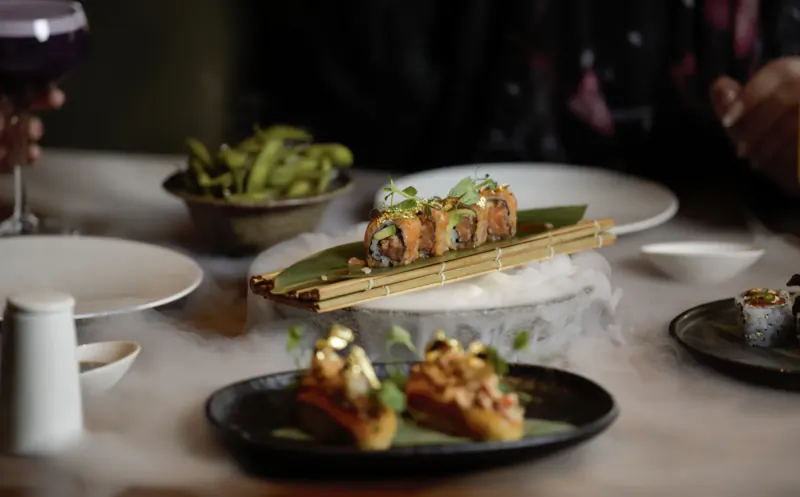 Assortment of sushi rolls, edamame, and topped baguette toasts on plates at Myazú Restaurant table with wine glasses.