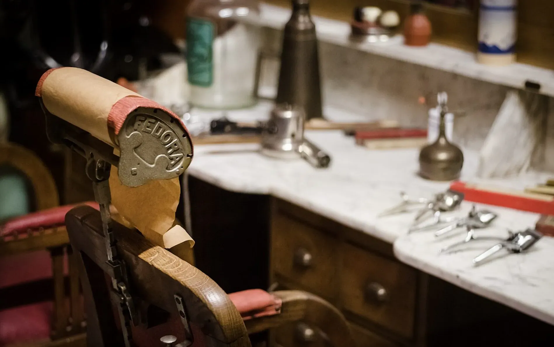Vintage barber chair with 'BARBER' medallion, facing marble counter with tools, razors, and bottles in shop.