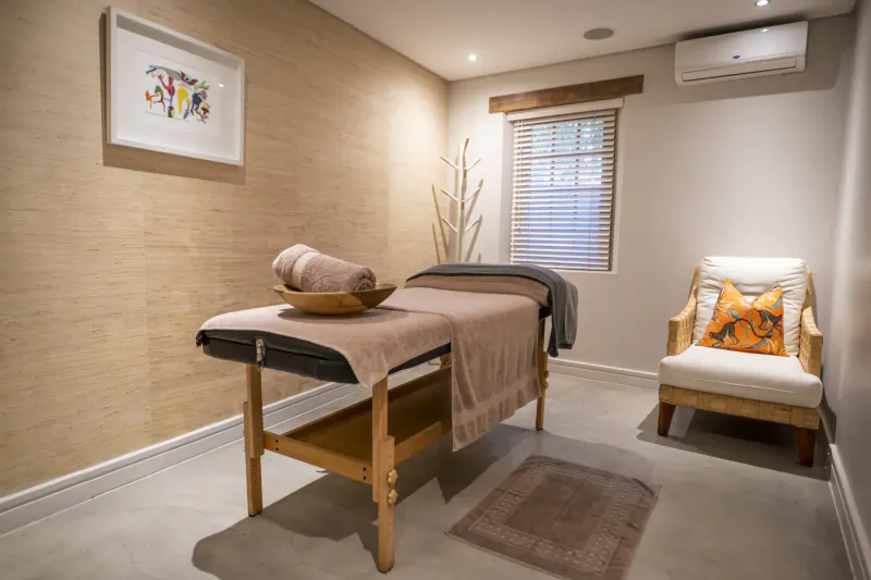 Cozy massage room with wooden massage table draped in towels, armchair, abstract wall art, window, and air conditioner.
