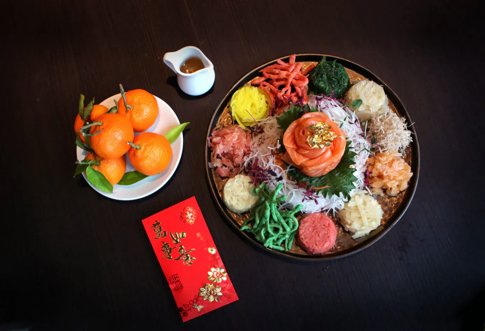 Vibrant yu sheng salad on plate with salmon rose, topped with orange slices, beside mandarin oranges, dipping sauce, and red packet on dark table.