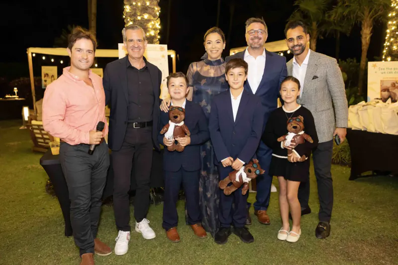 Group of adults and children holding teddy bears at Continuum childhood cancer society event on lawn at night.