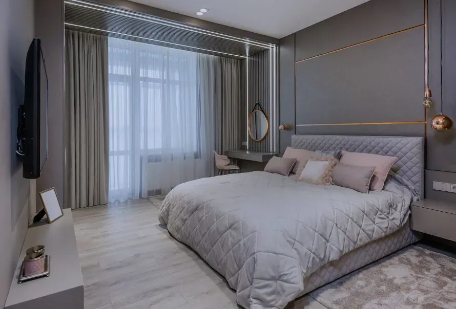 Luxurious modern bedroom with gray walls, king bed with pink pillows, vanity, TV, and large window drapes.