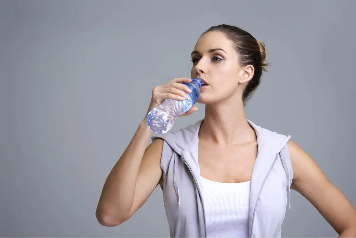 Fit woman in gray hoodie and tank top drinking from blue water bottle against gray background