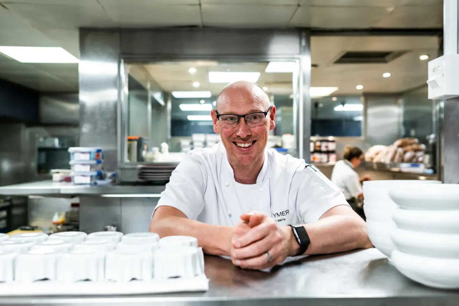 Steve Smith, head chef at Pennyhill Park, smiling in white chef uniform at stainless steel kitchen counter with stacked bowls