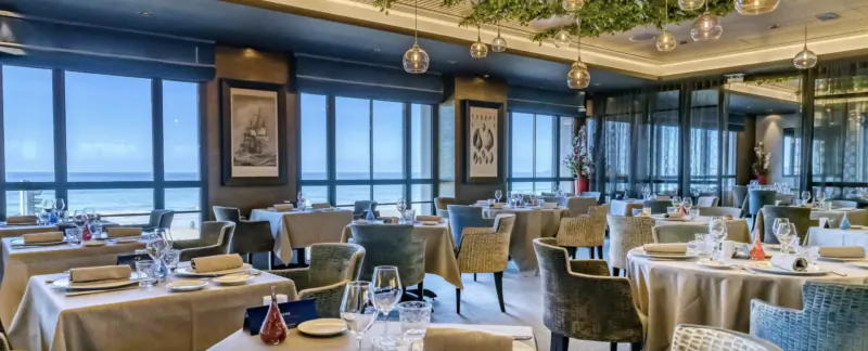 Elegant dining room in Saint-Malo with ocean views, white tablecloths, set tables, pendant lights, and framed art.