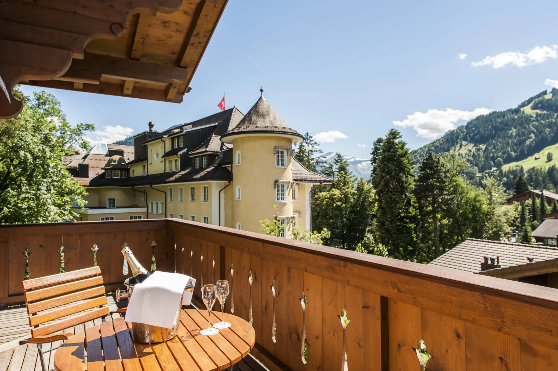 Wooden balcony with wine glasses and champagne on table, overlooking yellow chalet hotel and alpine mountains under blue sky.
