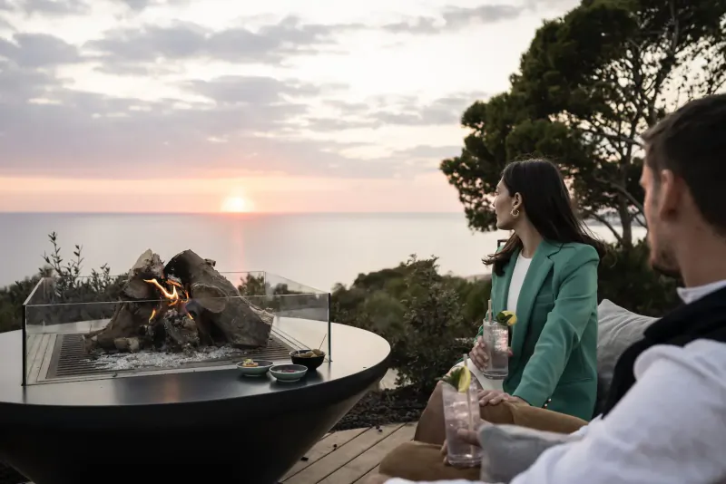 Couple in green blazer and white shirt relax with drinks by modern fire pit on deck, sunset over sea, ADLER Spa Resort Sicily