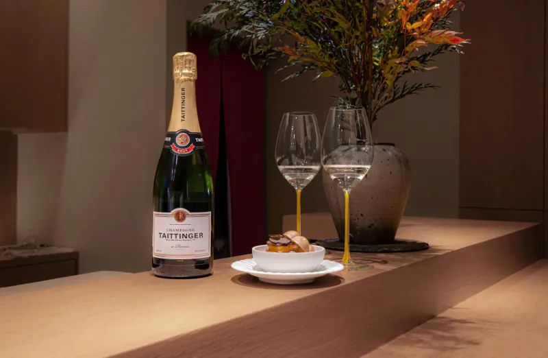 Taittinger champagne bottle, two tall glasses, and small white bowl with yellow garnish on wooden bar with plants.