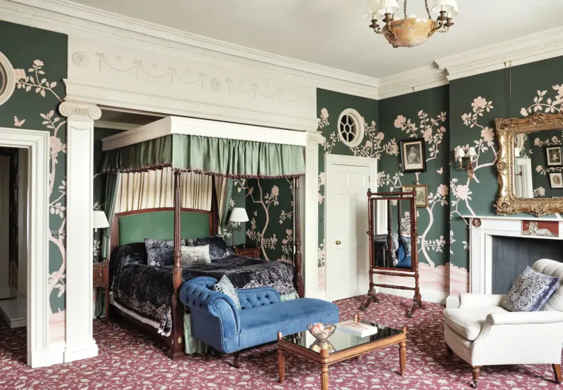 Elegant bedroom in Home House club with green floral wallpaper, four-poster bed, blue chaise, white armchair, and chandelier.