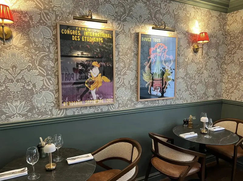 Elegant brasserie interior with green walls, ornate wallpaper, vintage French congress posters, and round tables set for dining.