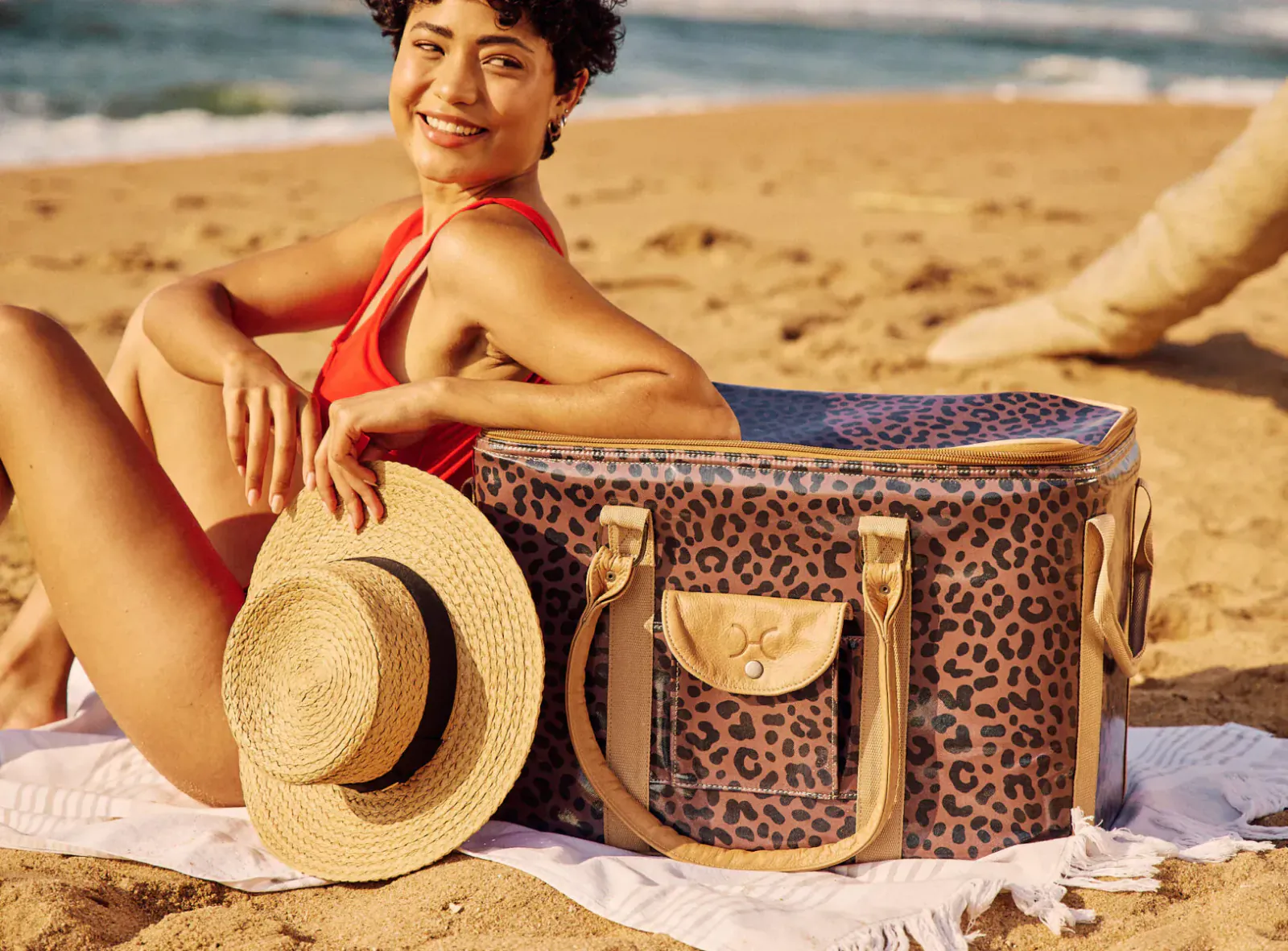 Smiling woman in red swimsuit on beach, posing with leopard-print cooler bag and straw hat