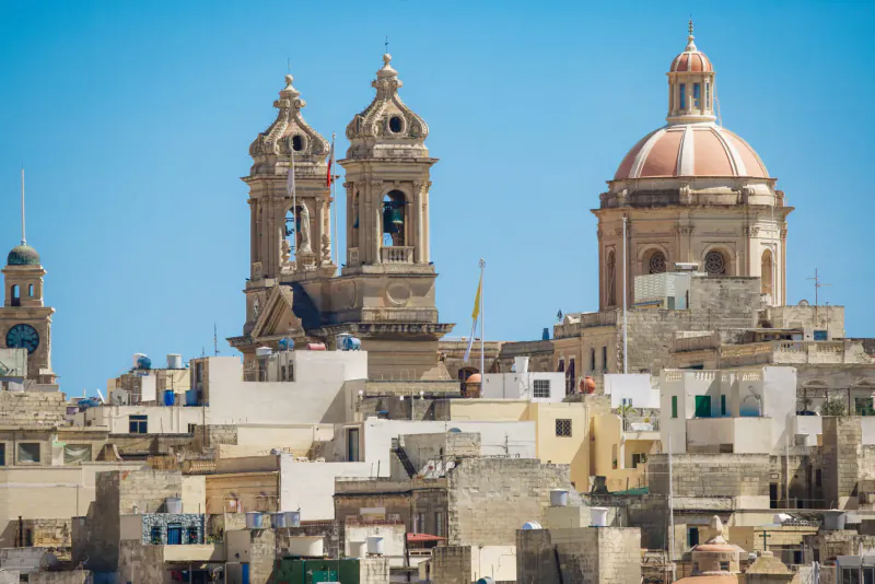 Panoramic view of twin-towered church with pink dome and Maltese village rooftops under blue sky