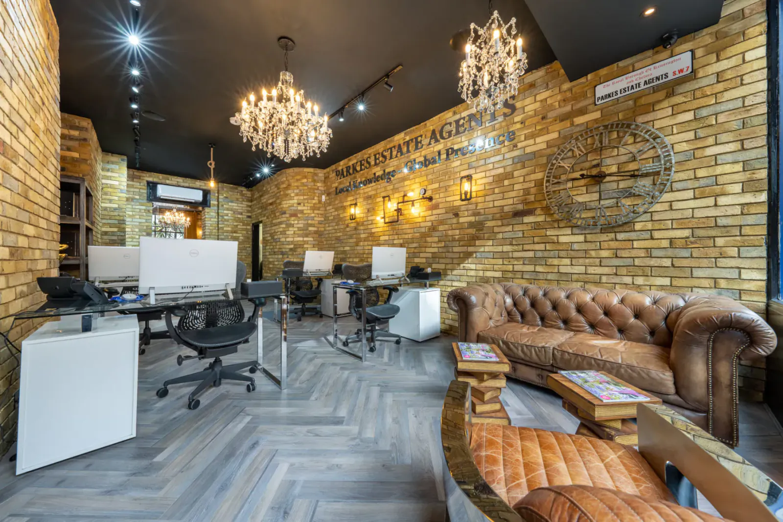 Modern Parkes Estate Agents office with brick walls, chandeliers, desks, leather sofa, and 'Parkes Estate Agents' sign.