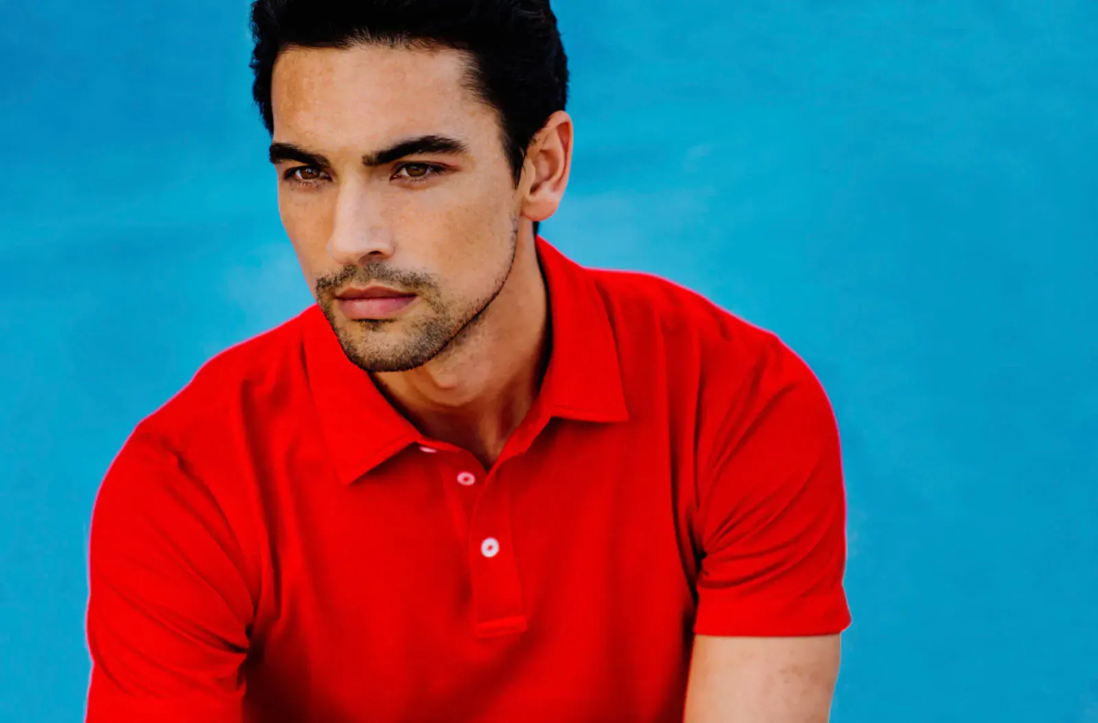 Handsome man with intense gaze wearing red polo shirt against blue background