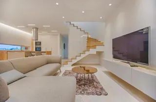 Modern lavish living room with beige L-shaped sofa, round wooden coffee table, white TV wall unit, open kitchen, and oak staircase