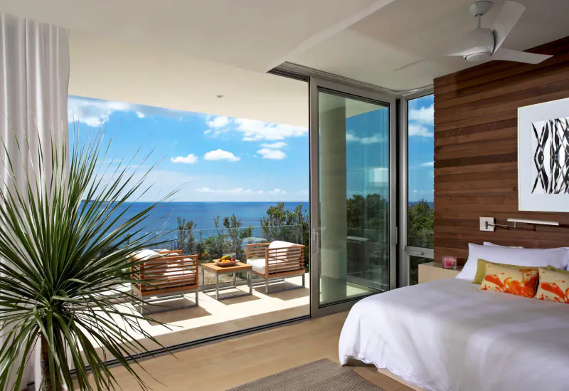 Modern bedroom in private resort with sliding doors to balcony overlooking ocean, lounge chairs, plants, and artwork.