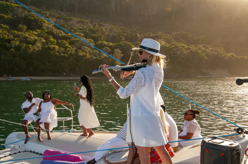 Blonde woman in white dress and hat playing violin on yacht deck with group in white at sunset by ocean, Cape sailing charter.