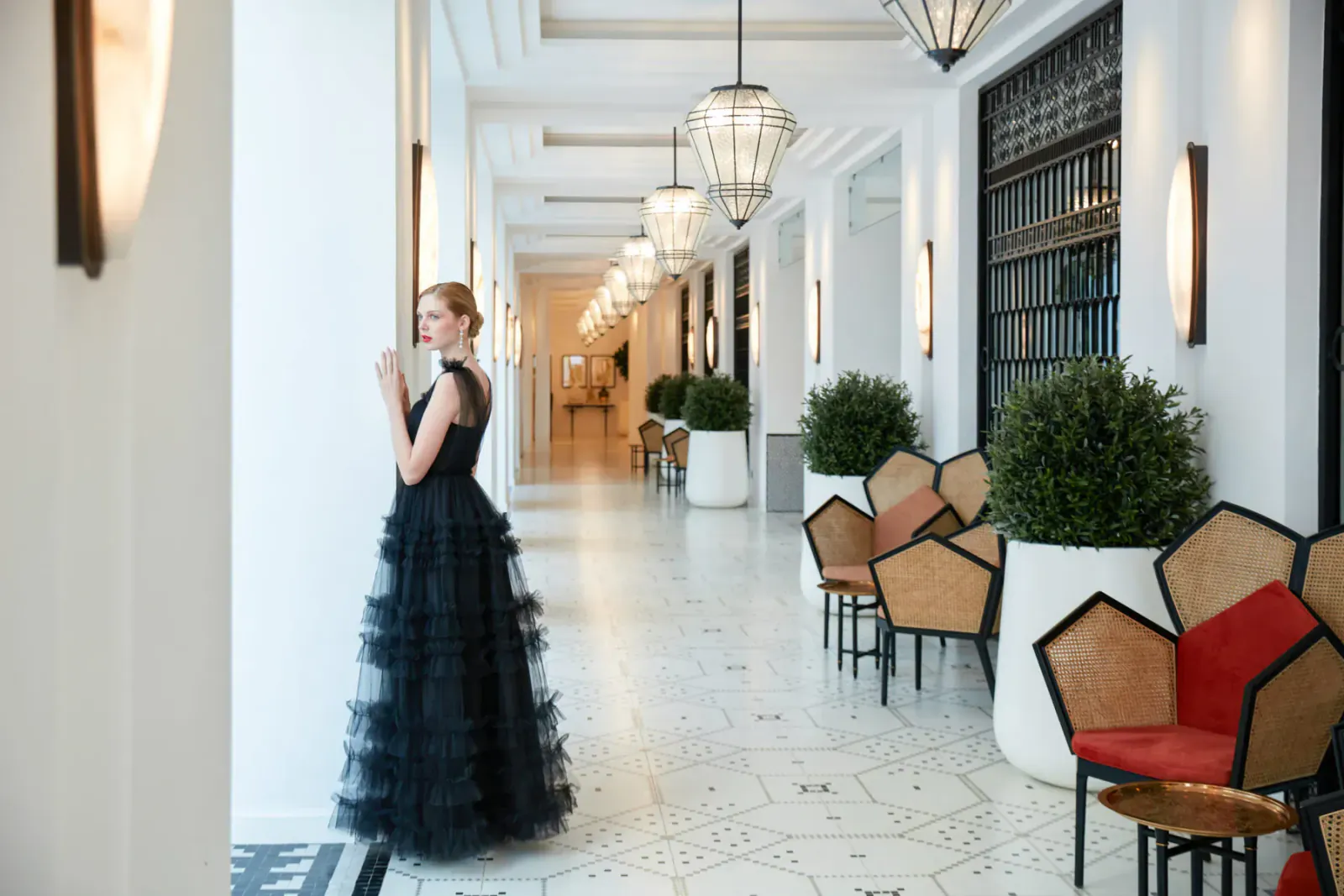 Blonde woman in black tulle gown stands in elegant colonnaded hotel hallway with pendant lights, rattan chairs, and plants