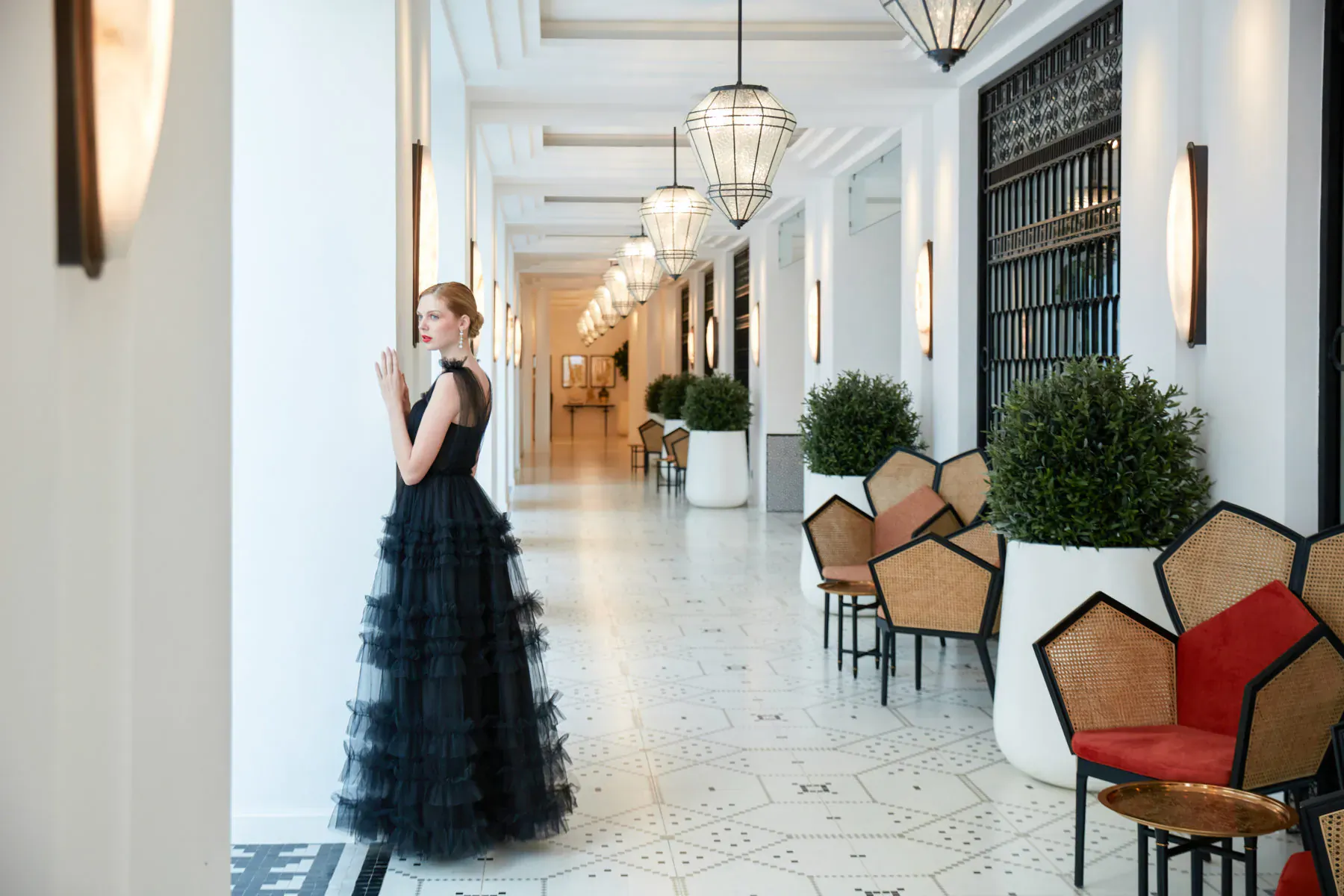 Blonde woman in black tulle gown stands in elegant colonnaded hotel hallway with pendant lights, rattan chairs, and plants