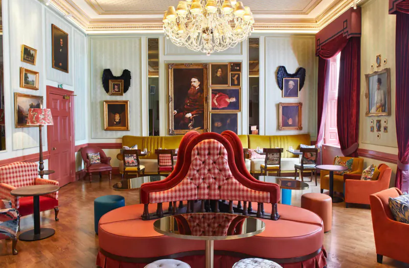 Opulent Home House gallery lounge with chandelier, large portrait, red tufted seating, and eclectic art on pale walls.