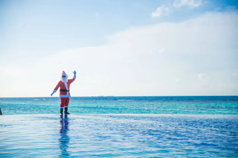 Santa Claus in red suit waving on infinity pool edge overlooking turquoise ocean at beach resort