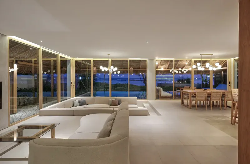 Spacious living room in 3-bedroom beachfront pool villa at Amber Lombok, with white L-shaped sofa, wood dining table, large windows to pool at dusk.