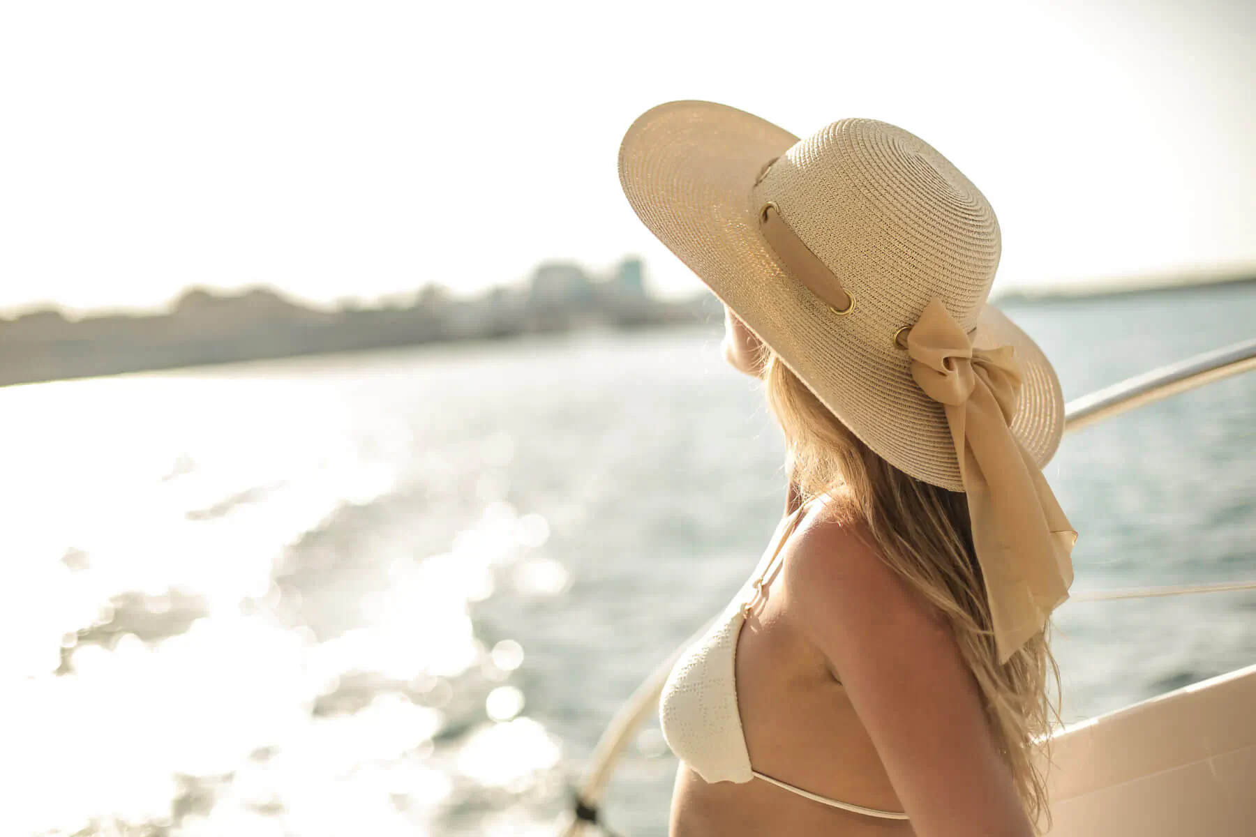 Blonde woman in white bikini and wide-brim hat stands on luxury yacht, gazing at sunlit ocean horizon