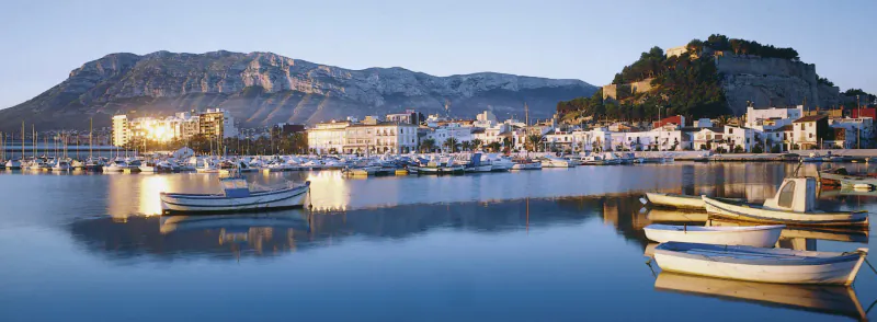 Panoramic view of a marina with white boats reflected in calm water at dusk, backed by a cliffside town and mountain.