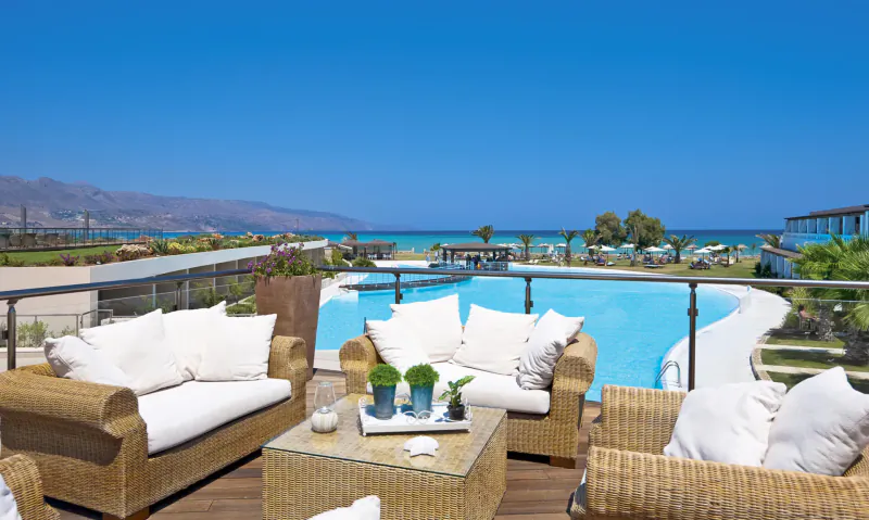 Luxurious outdoor lounge with white cushions on wicker furniture, overlooking infinity pool, sea, and mountains at Cavo Spada resort in Crete.