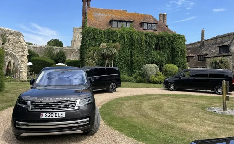 Black Range Rover (52E LE) parked on gravel driveway amid manicured lawns before ivy-covered historic castle with black chauffeur vans nearby.