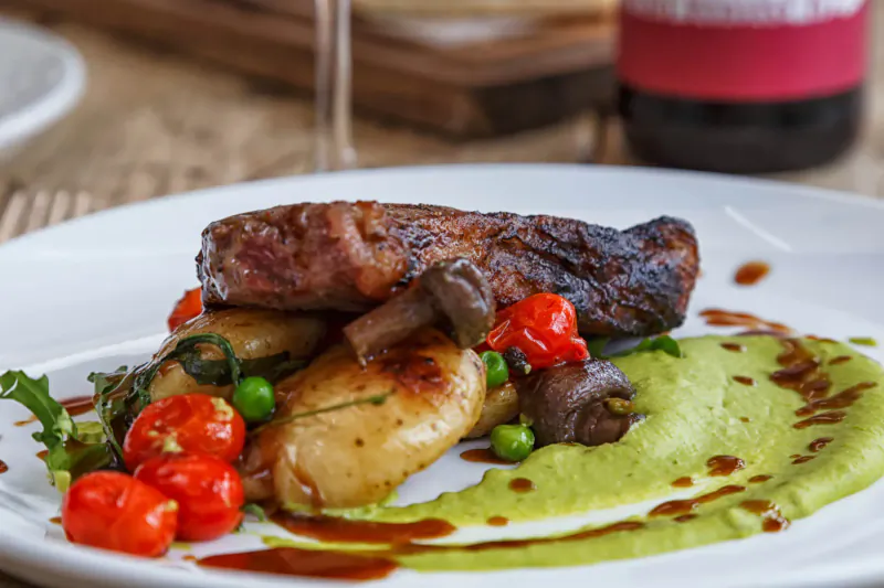Grilled sausage with mushrooms, roasted potatoes, peas, cherry tomatoes, and green pea puree on white plate