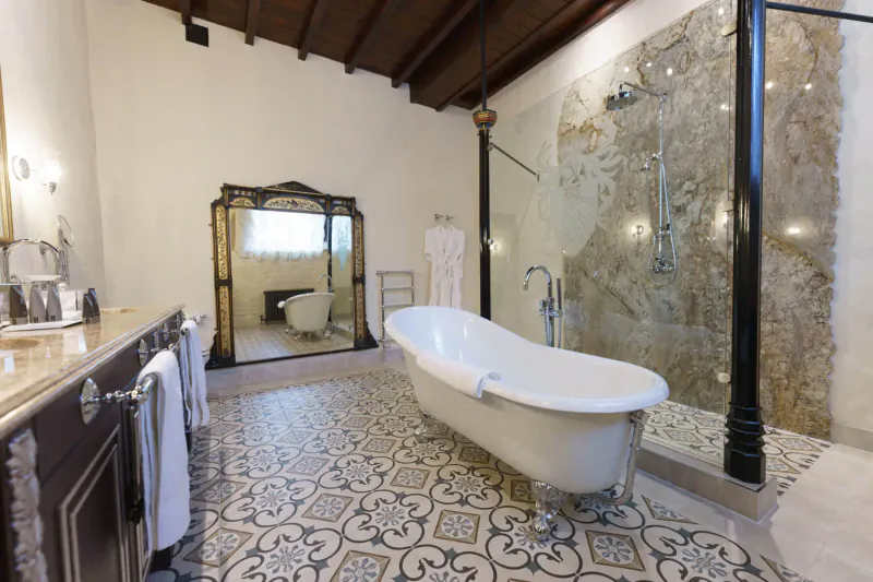 Elegant Thornbury Castle bathroom with freestanding white bathtub, ornate mirror, stone shower, and tiled floor.