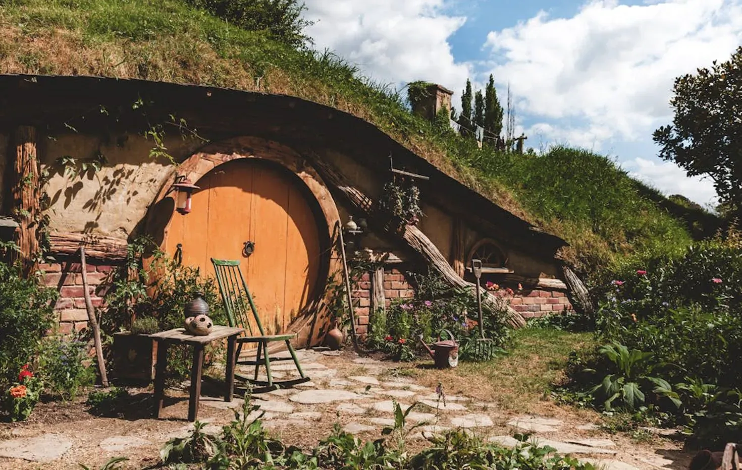 Hobbit house with round orange door, grass roof, garden, chair, and table in lush yard under blue sky