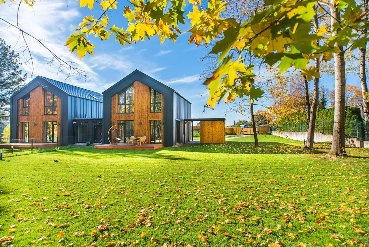 Two modern wooden custom outbuildings with black accents in lush green yard, framed by autumn yellow trees under blue sky.