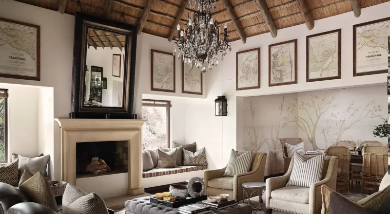 Luxurious savanna lodge living room with thatched ceiling, chandelier, fireplace, plush chairs, and framed maps.