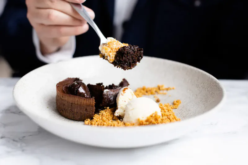 Hand with spoon lifting bite of chocolate torte topped with ice cream and crumble from white plate