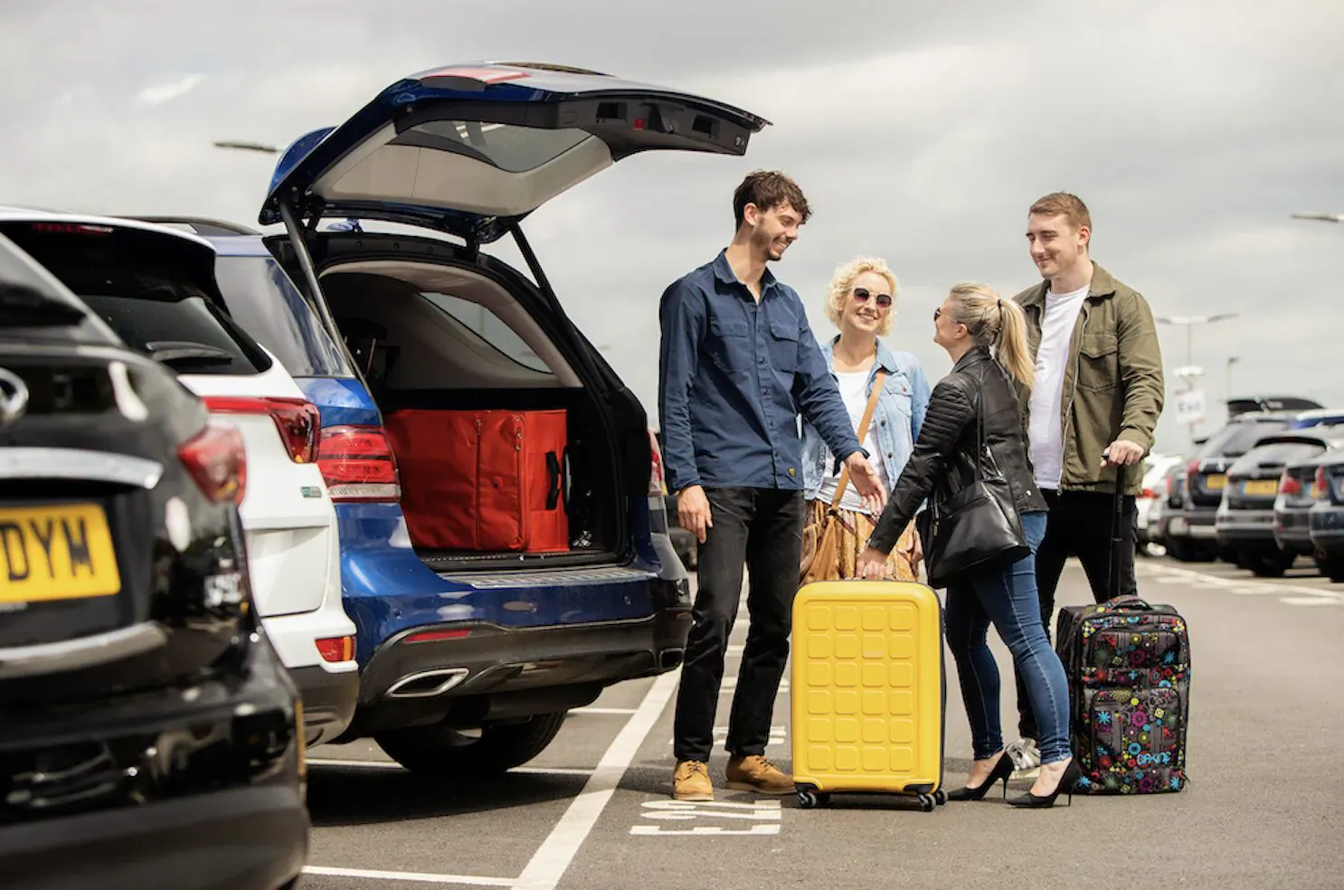 Four young adults with colorful suitcases at open blue Nissan SUV boot in Gatwick Airport parking