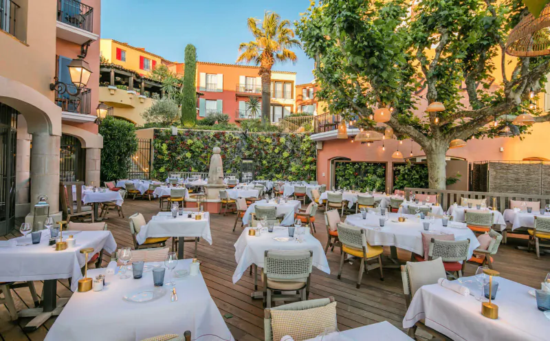 Elegant outdoor terrace at Byblos Palace hotel in Saint-Tropez with white-clothed tables, palm trees, fountain, and sunset views.