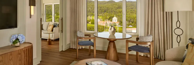 Luxurious hotel suite with blue armchairs at round wooden table, hydrangeas, sofa, and panoramic view of green hills and dome from large windows.