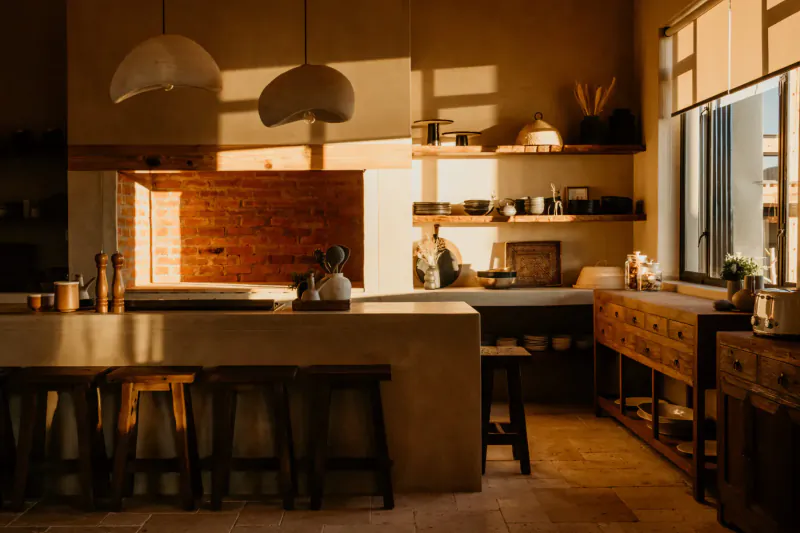 Warmly lit modern kitchen in Savanna Luxury Villa, Namibia, with wooden stools at stone island, brick backsplash, ceramics on shelves, golden sunlight.