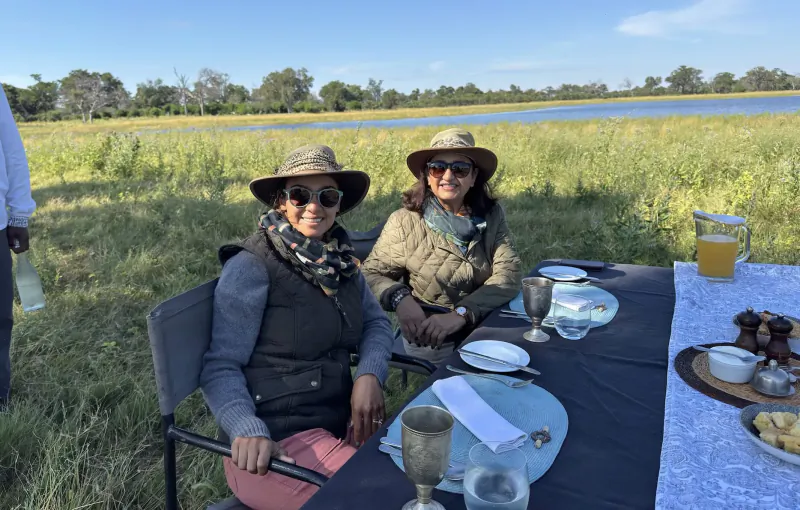 Two women in hats and jackets seated at an outdoor table with wine glasses, juice pitcher, and snacks by a lagoon and grassland.