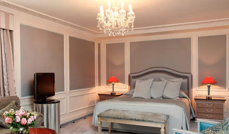 Elegant Bentley Hotel suite in Kensington with chandelier-lit gray king bed, red lamps, TV, flowers.