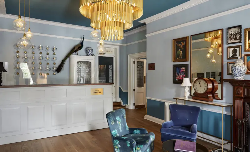 Elegant hotel reception with white desk, gold chandelier, peacock decor, vintage clock on mantel, blue armchairs