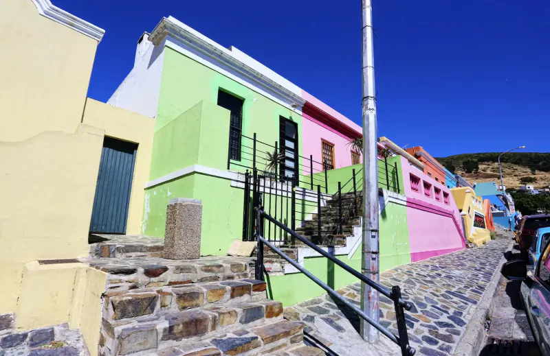 Colorful Cape Malay houses in vibrant pinks, greens, and yellows line a cobbled street under blue sky.