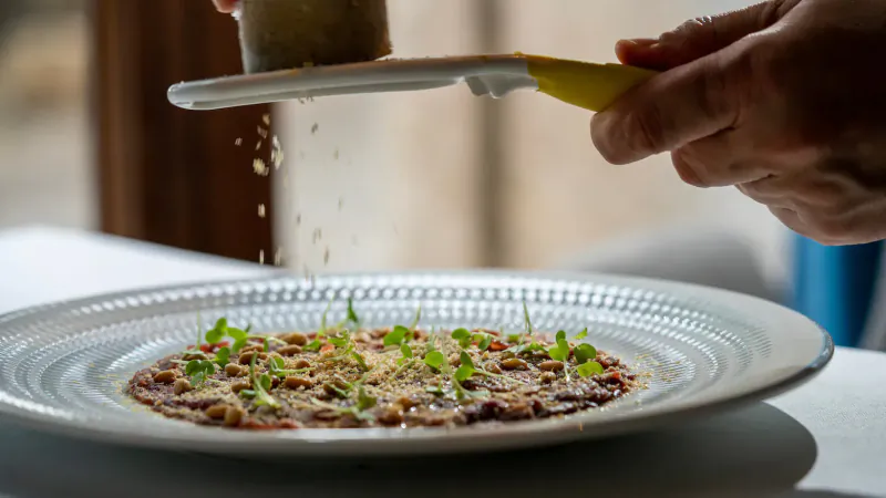 Hand grating cheese over plated dish with beans, microgreens, and red chilies at El Restaurant El Llorenç.